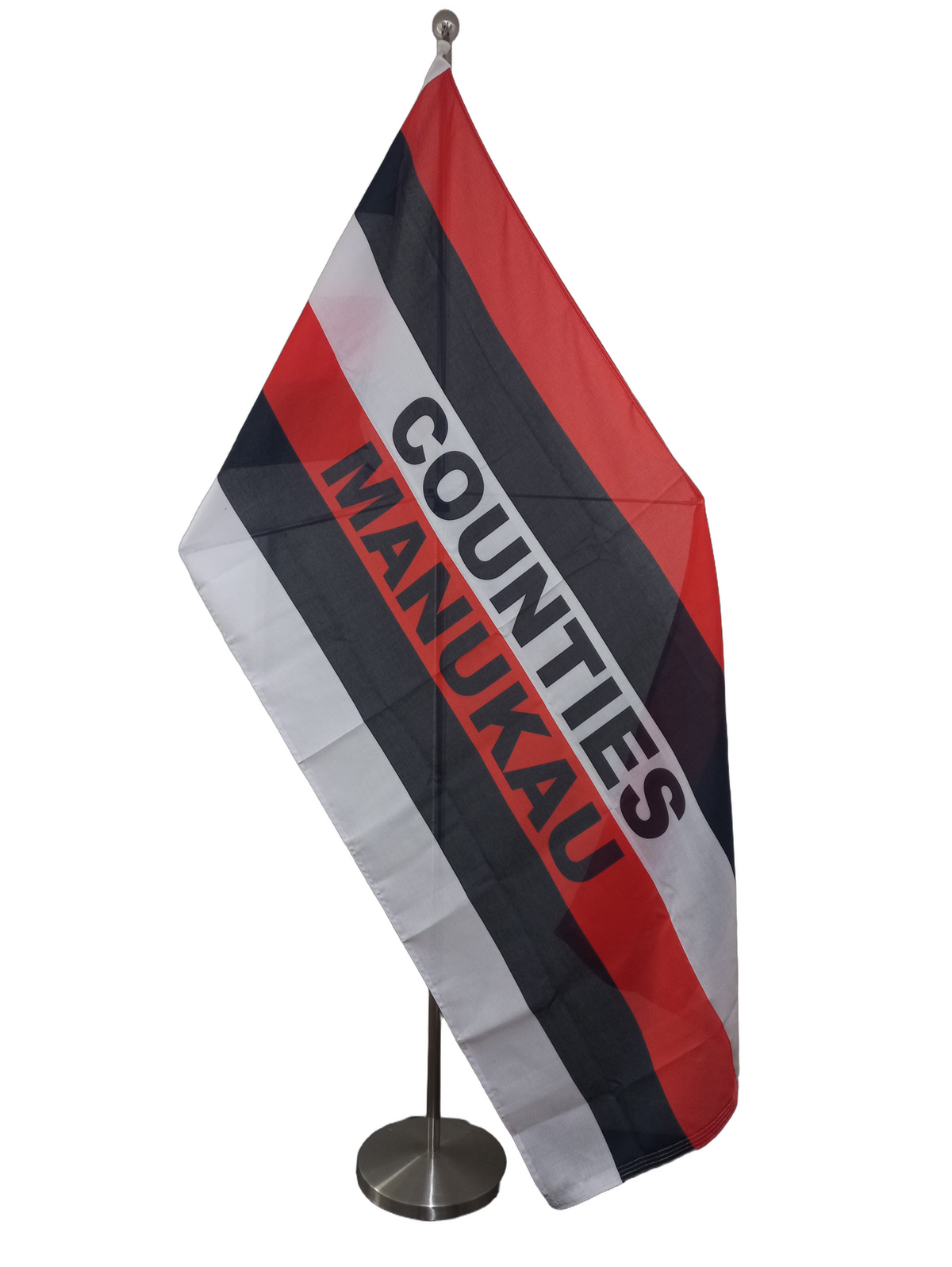A rectangular flag with horizontal red, black and white stripes and the text 'COUNTIES MANUKAU' in black. On a ceremonial flag pole.